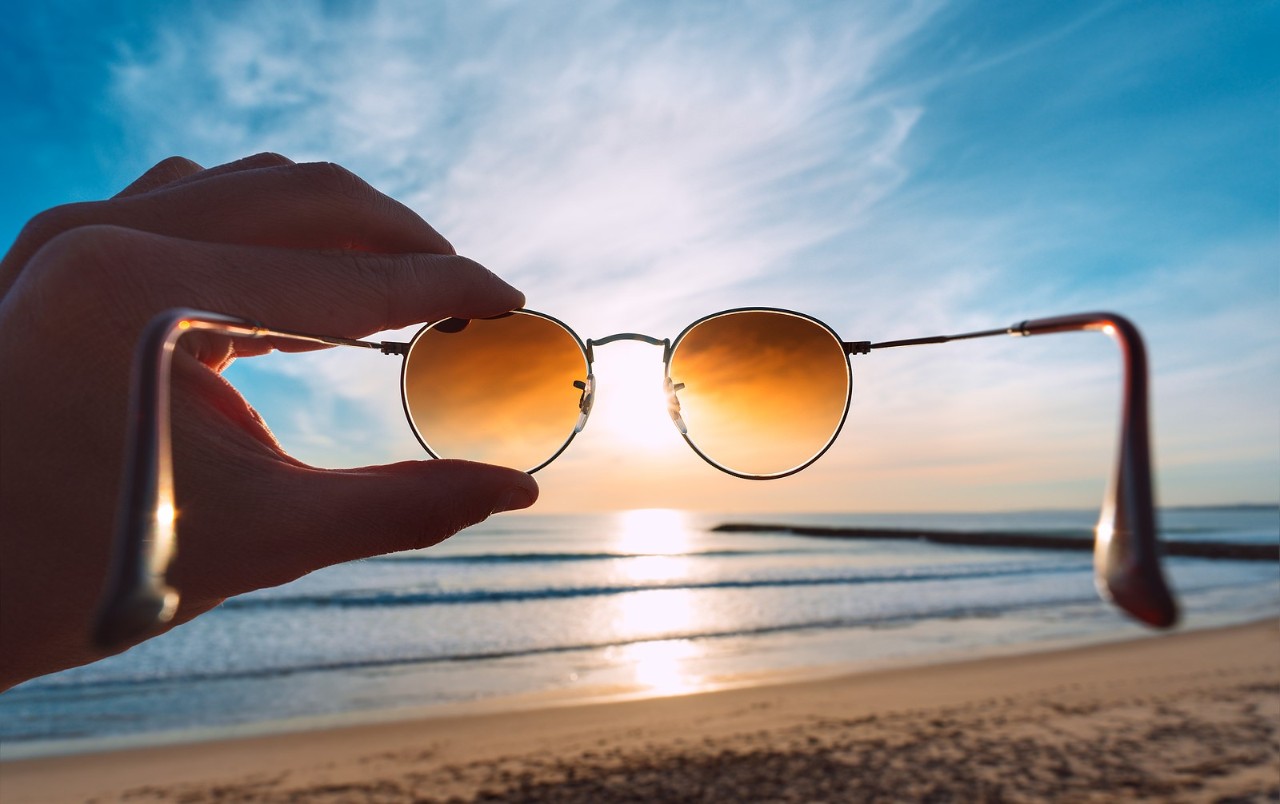 Hand holding stylish round sunglasses with brown lenses at sunset. Putting on sunglasses at sunny summer day near the ocean. Man looking at bright sun through polarized sunglasses. Summer vibes